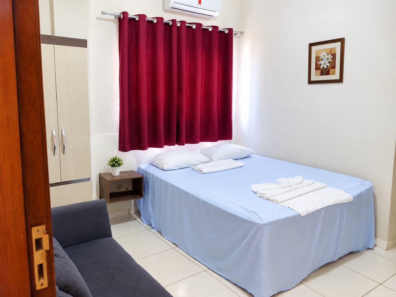 Apartamento casal com cama arrumada, toalhas, cortina vermelha, ar-condicionado e sofá
